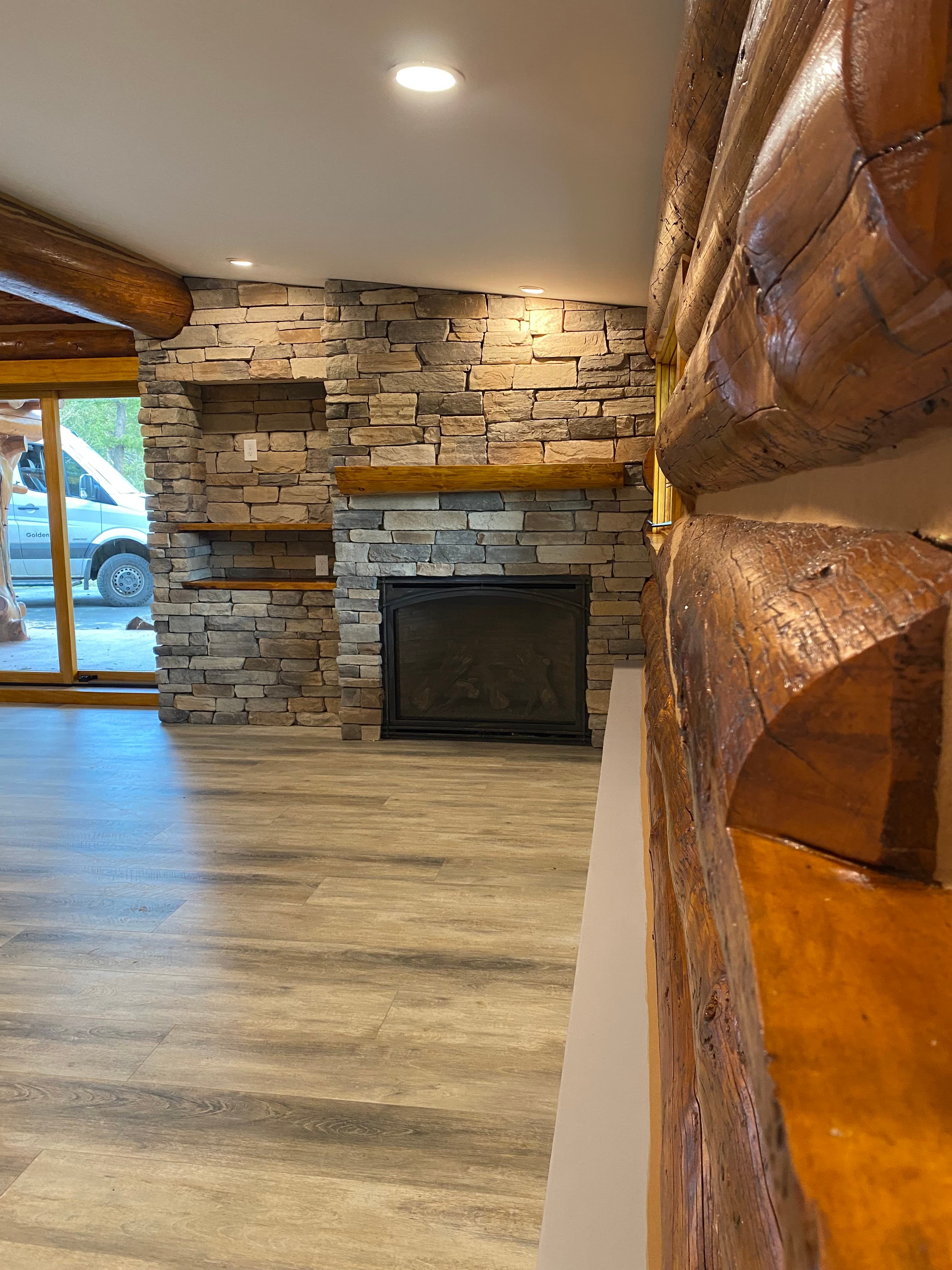 Stacked stone fireplace with a black arched insert, a log mantel, and built-in wood shelves in a log cabin