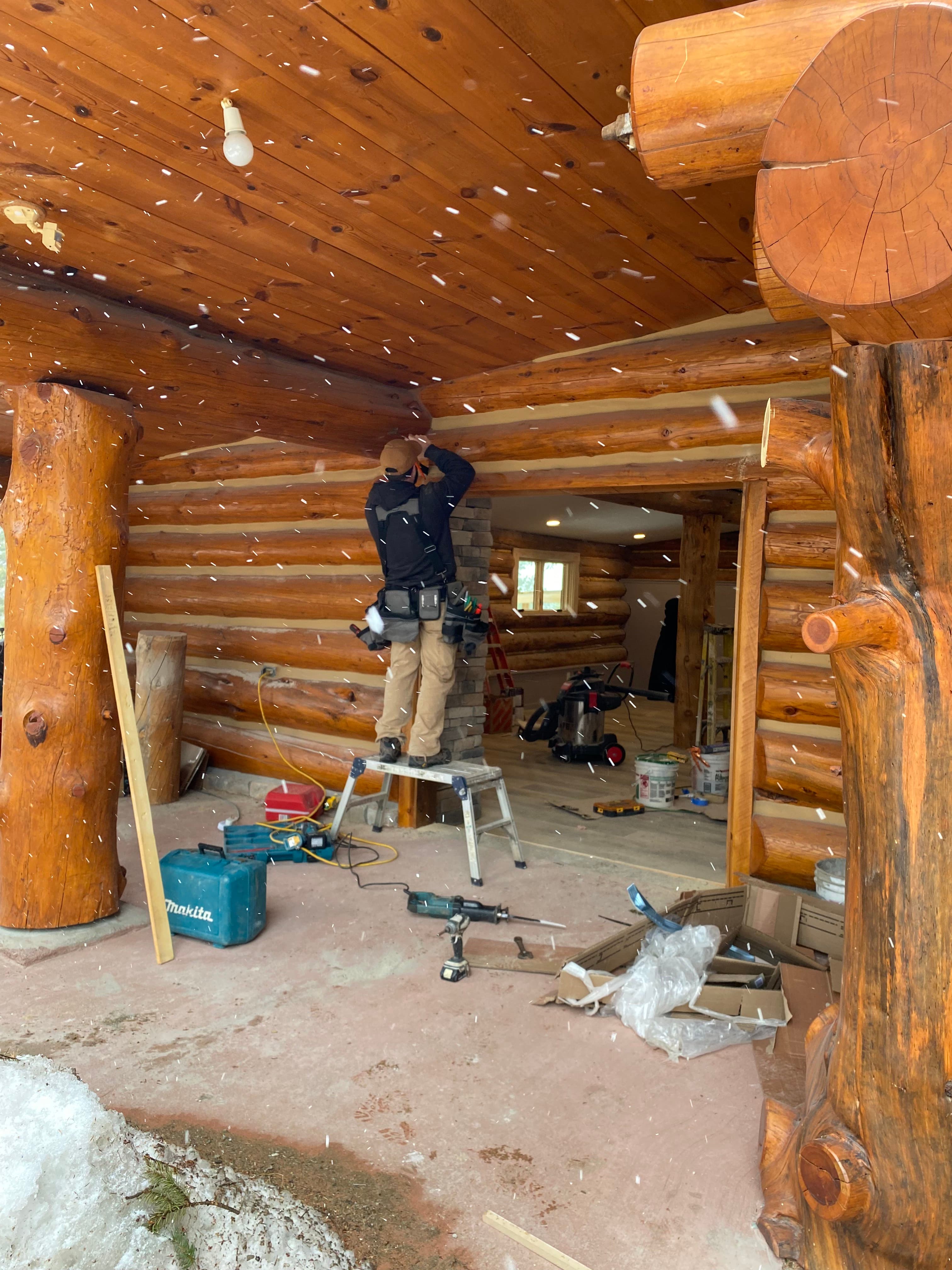 Work on a log cabin porch in falling snow, with tools on the floor and the interior fireplace visible through the door